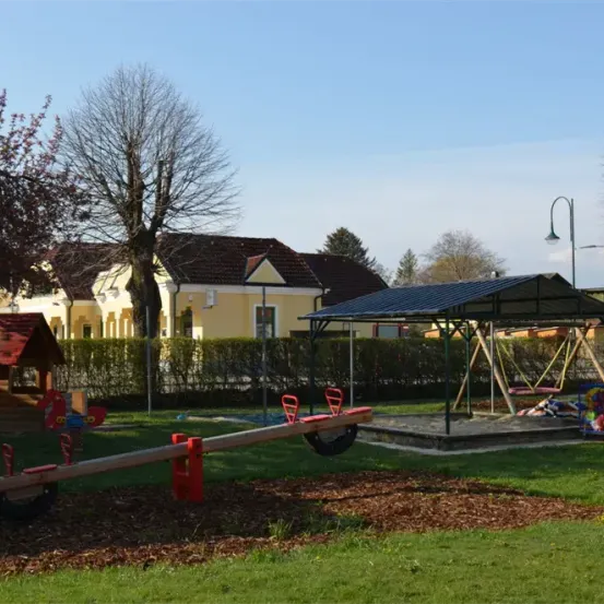 Ein Spielplatz mit einer Rutsche und einem überdachten Pavillon. Dahinter ein gelbes Haus mit einem braunen Dach. Das Gelände ist von Büschen und Bäumen umgeben.