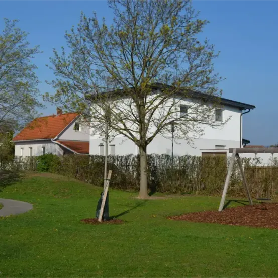 Ein weißes Haus mit einem roten Dach steht hinter einem Baum und einer Hecke. Ein Schaukel ist in einem Hof platziert. Der Himmel ist klar.