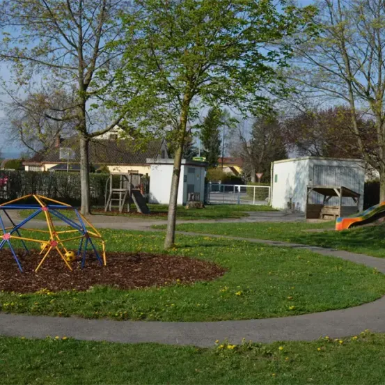 Ein Spielplatz mit einer bunten Kuppel, einer Rutsche und einem Schaukelset. Der Bereich ist von Bäumen und Gras umgeben. Im Hintergrund steht ein Haus.