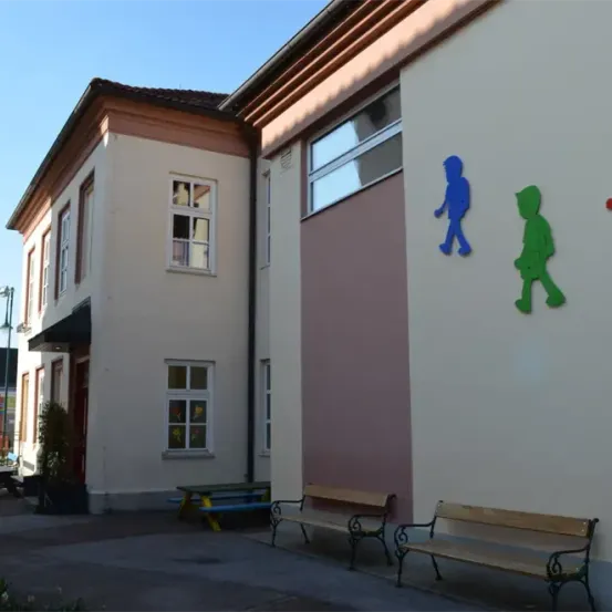 Ein weißes Gebäude mit Fenstern und einem roten Dach hat eine Bank davor. An der Wand befinden sich drei bunte Figuren, ein blauer Junge, ein grüner Junge und ein rotes Mädchen.