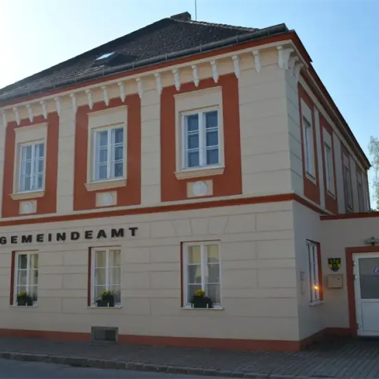 Ein zweistöckiges Gebäude mit weißen Wänden und orangefarbenem Rand. Das Schild steht 'Gemeindeamt'. Es gibt sechs Fenster mit Pflanzen in den Fensterbänken.