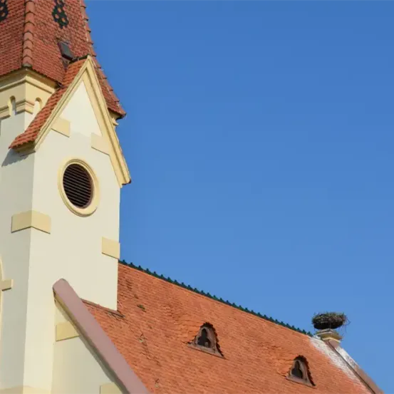 Das Bild zeigt einen Kirchturm mit einer Uhr und einem Vogelnest auf seinem Dach. Der Turm ist in einer hellen Farbe, möglicherweise weiß, mit einem roten Dach gestrichen. Die Uhr befindet sich auf der Vorderseite des Turms.