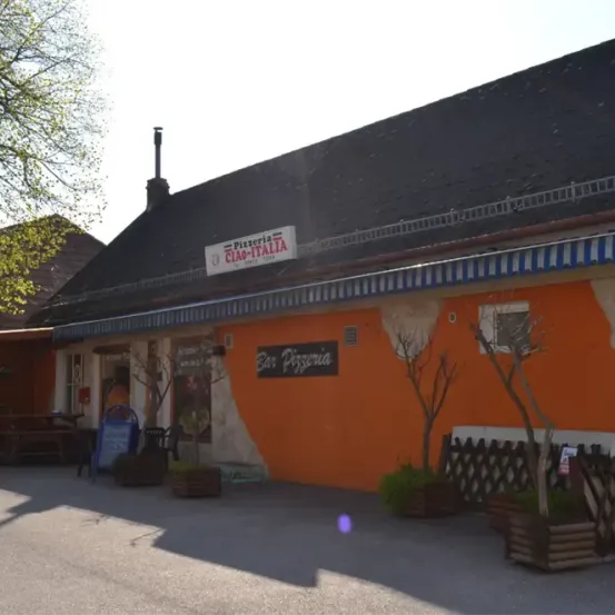 Ein orangefarbenes Gebäude mit einem Schild, auf dem 'Pizzeria CIAO-ITALIA' steht. Das Gebäude hat ein blau-weiß gestreiftes Vordach. Stühle und ein Tisch sind draußen aufgestellt. Vor dem Gebäude stehen Topfpflanzen und ein Holzzaun.