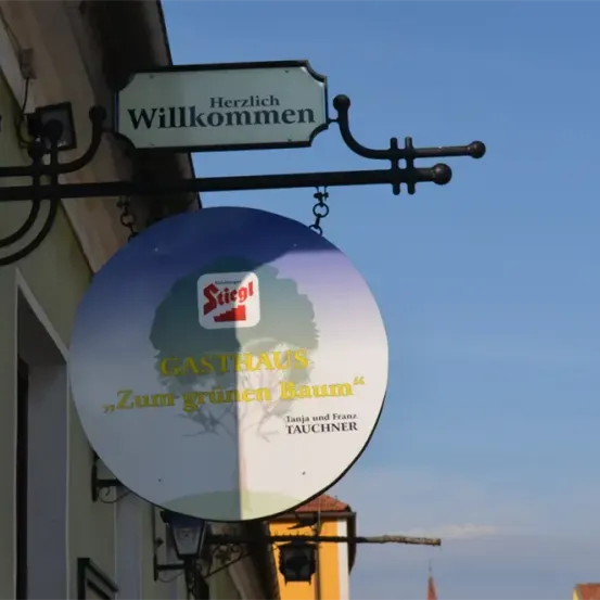 Ein Schild für Gasthaus Zum grünen Baum hängt an einem Gebäude. Das Schild ist blau mit einem Baumlogo und Text auf Deutsch.