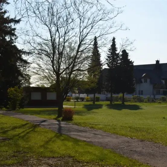 Ein Rasenpark mit einem Weg, der zu einem Haus mit schwarzem Dach und weißen Wänden führt. Der Bereich ist von Bäumen und Sträuchern umgeben.