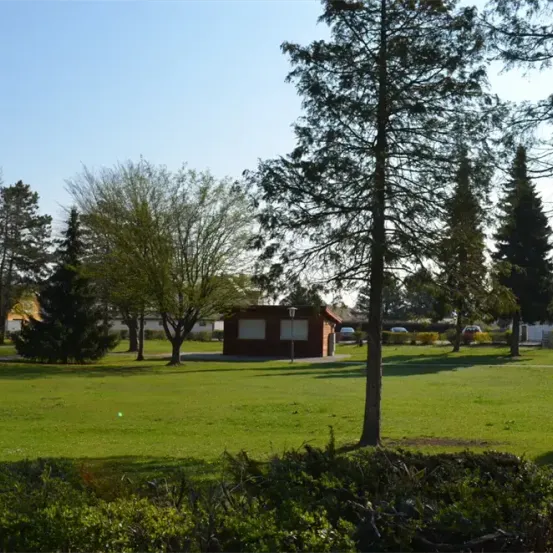 Ein Park mit saftigem grünem Gras, Bäumen und einem kleinen Gebäude. Der Himmel ist klar und blau.
