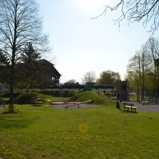 Ein Kinderspielplatz mit Rutsche, kleinem Hügel und Bänken in einem Park mit einem Haus und Bäumen im Hintergrund.