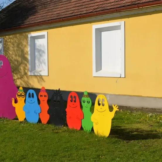 Eine Gruppe farbenfroher, lächelnder Cartoon-Charaktere steht vor einem gelben Haus mit weißen Fenstern.