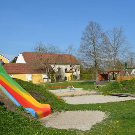 Ein bunter Rutschen- und Spielbereich vor einem Haus mit rotem Dach, einem Holzschuppen und Bäumen. Der Himmel ist klar und blau.