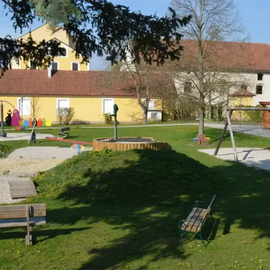 Ein Spielplatz mit einem roten Dachgebäude im Hintergrund. Bänke und Schaukeln sind im Vordergrund.