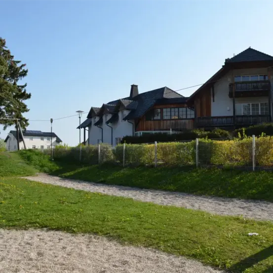 Ein Feldweg führt zu einer Reihe von Häusern unter einem klaren blauen Himmel. Die Häuser haben Holzakzente und sind von Grünflächen und einem Zaun umgeben.