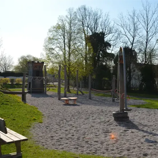 Freiluftspielplatz mit einem Holzturm, einer Schaukel und einem Rutschen, umgeben von Bäumen. Eine Bank steht in der Nähe des Sandbereichs.