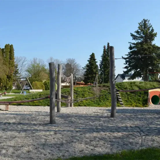 Ein Outdoor-Spielplatz mit Holzpfosten, einem Seil und einer Rutsche. Im Hintergrund befinden sich Bäume und Häuser.