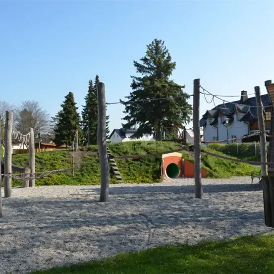 Ein Outdoor-Spielplatz mit verschiedenen Kletterstrukturen, einem Tunnel und einer Rutsche, vor dem Hintergrund von Grünflächen und Häusern unter einem klaren Himmel.