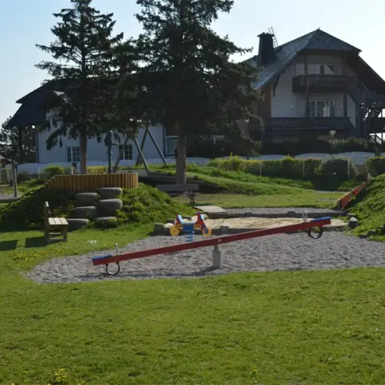 Ein Spielplatz mit einer Wippe und einem kleinen roten Fahrrad auf dem Boden. Bäume und ein weißes Haus im Hintergrund.