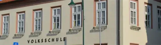 Ein beiges Gebäude mit vielen Fenstern und Balkonen, beschriftet mit 'Volksschule'. Ein Straßenlicht steht davor.