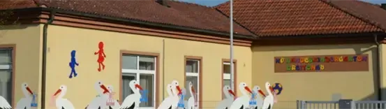 Ein Gebäude mit einem braunen Dach hat viele Fenster und ein Schild. Es gibt mehrere Störche, die auf der Wand gemalt sind.