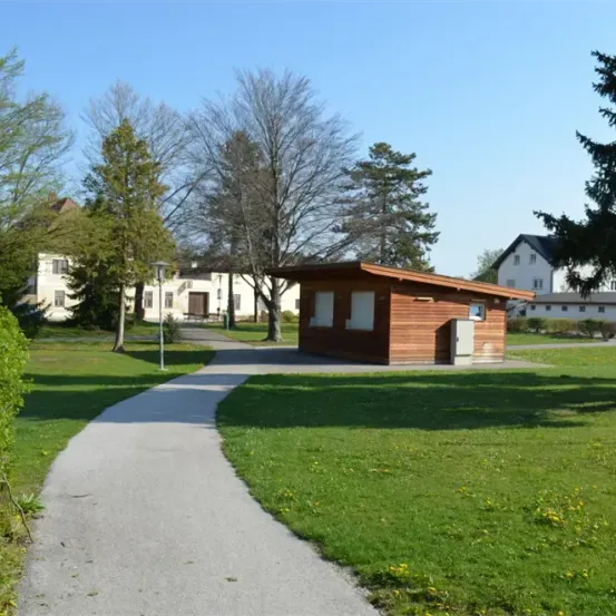 Ein Weg führt zu einem kleinen Holzgebäude in einem Park mit üppigem grünem Gras und Bäumen unter einem klaren blauen Himmel.