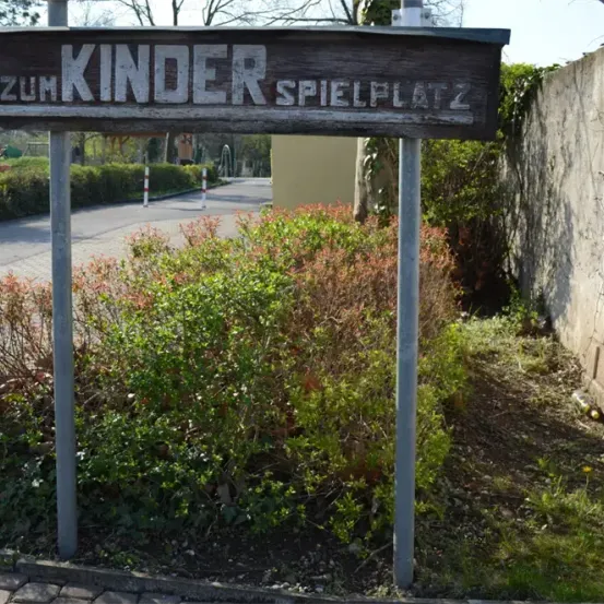 Ein hölzernes Schild mit weißen Buchstaben steht am Straßenrand. Es steht 'Zum Kinder Spielplatz' und zeigt auf einen Spielplatz hin.