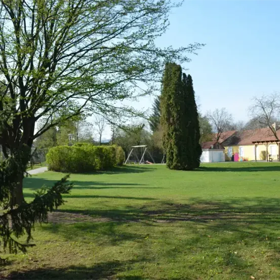 Ein Park mit einem Rasenfeld, Büschen, einem Baum und einem Haus mit braunem Dach. Es gibt eine Schaukel und eine Bank. Der Himmel ist klar und blau.