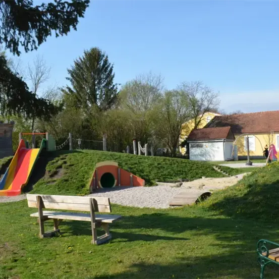 Ein Spielplatz mit einer bunten Rutsche, einer Holzbank und einem kleinen Tunnel an einem sonnigen Tag. Im Hintergrund sind Bäume und Häuser zu sehen.