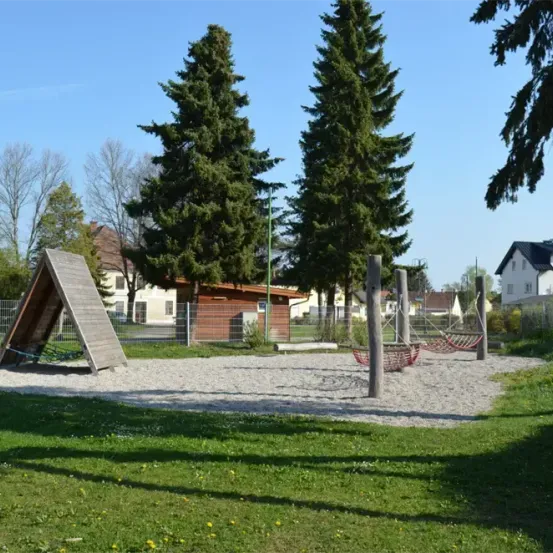 Ein Spielplatz mit einem roten Schaukel, einer dreieckigen Holzstruktur und zwei roten Schaukeln. Bäume und Häuser im Hintergrund. Der Boden ist mit Kies bedeckt.