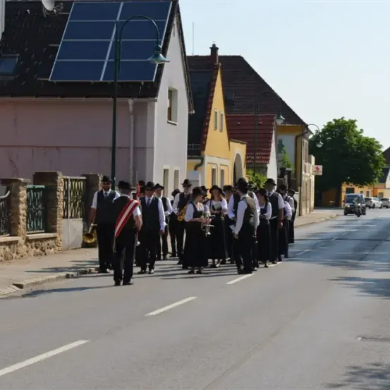 Eine Gruppe von Menschen in traditioneller Kleidung marschiert auf der Straße, einige halten Musikinstrumente. Hinter ihnen sind Häuser mit Solarpanelen und einige Autos zu sehen.
