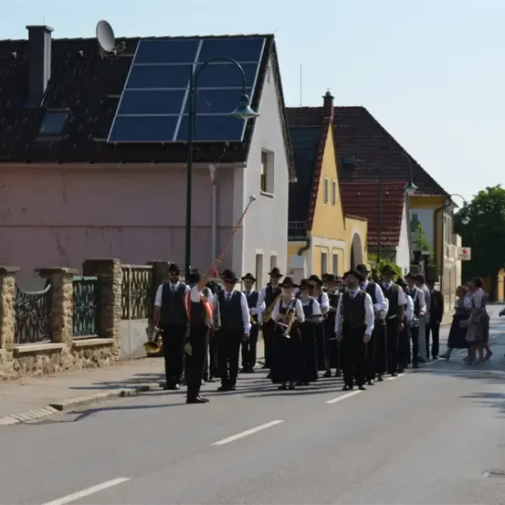 Eine Blaskapelle spielt auf einer Straße in einer Kleinstadt, mit Häusern und Solarmodulen im Hintergrund.