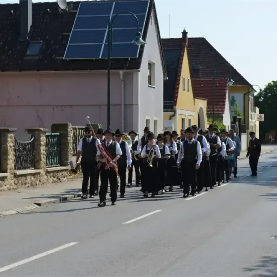 Eine Blaskapelle in traditioneller Kleidung marschiert in einem Dorf die Straße entlang. Sie spielen Trompeten und Posaunen.