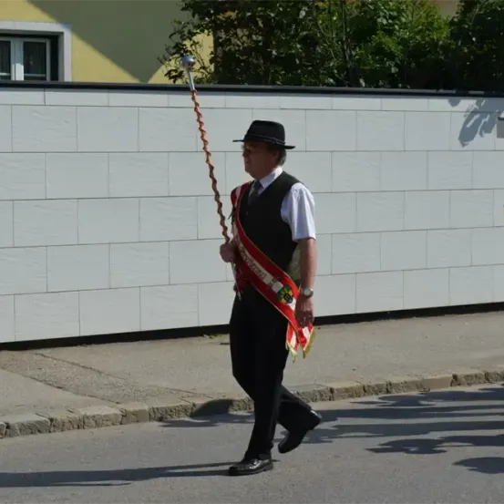 Ein Mann marschiert die Straße entlang, hält einen zeremoniellen Stab, trägt einen schwarzen Hut, eine Weste und schwarze Hosen, mit einer roten Schärpe über der Schulter. Hinter ihm spielt eine weitere Person ein Blechinstrument.