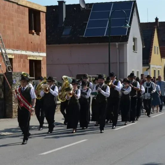 Eine Blaskapelle in traditionellen Uniformen, die in einer Stadtstraße marschiert, mit Zuschauern, die hinter ihnen gehen, Gebäude mit Solarpanelen im Hintergrund.