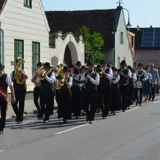 Eine Brassband in traditioneller Kleidung marschiert die Straße entlang, gefolgt von Schaulustigen. Gebäude säumen die Straße.