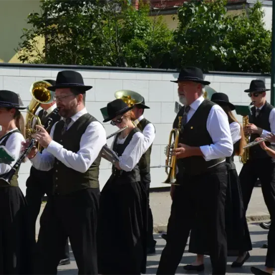 Eine Blaskapelle spielt Instrumente bei einem Umzug, mit Menschen in schwarzen Kleidern und Hüten. Sie sind aufgereiht und halten Musikinstrumente.
