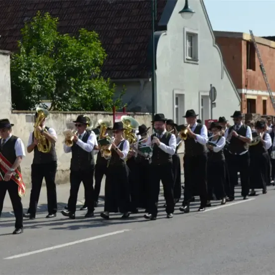Eine Blaskapelle mit verschiedenen Blechblasinstrumenten zieht die Straße entlang, an der Gebäude stehen.