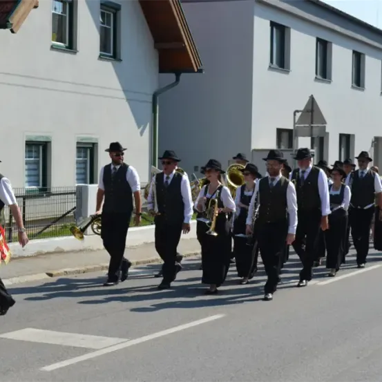 Eine Blaskapelle von Erwachsenen in formeller Kleidung geht vor einem Gebäude die Straße hinunter.
