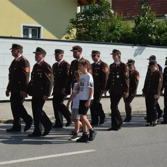 Eine Gruppe uniformierter Personen, möglicherweise Militär, marschiert die Straße entlang, begleitet von einem Jungen in einem weißen T-Shirt.
