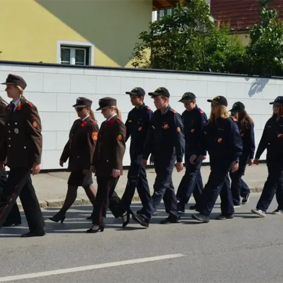 Eine Gruppe uniformierter Personen, wahrscheinlich Polizisten, marschiert die Straße entlang. Sie sind in formeller Kleidung und die Straße hat weiße Markierungen. Bäume und ein Gebäude sind im Hintergrund.