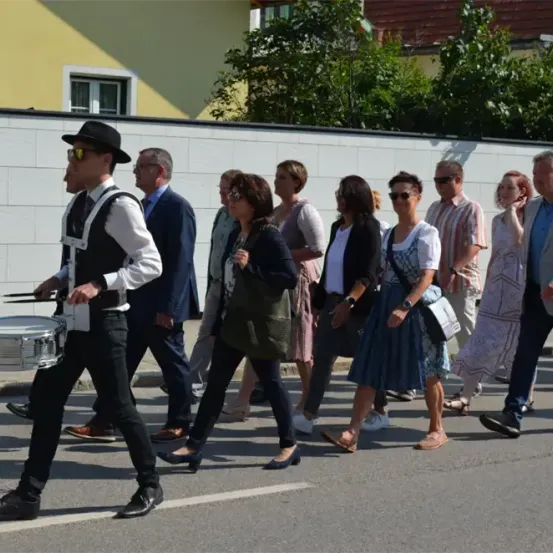 Eine Gruppe von Menschen marschiert in einer Parade, angeführt von einem Trommler in Anzug und Hut. Die Marschierer umfassen Männer und Frauen, alle in lässiger Kleidung. Sie gehen eine Straße entlang, gesäumt von einer weißen Mauer und Grünflächen.