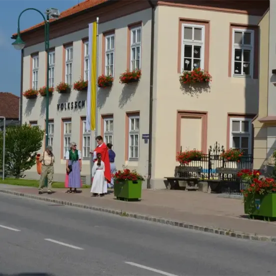 Ein Gebäude mit einem Schild, das 'VOLKSSCH' liest, hat Blumen an den Fenstern. Auf dem Bürgersteig steht eine Gruppe von Menschen in traditioneller Kleidung.