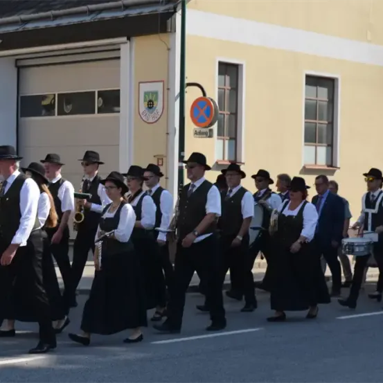 Eine Musikkapelle mit Bläsern und Schlagzeugern geht eine Straße entlang, vor einem Gebäude mit einem roten Schild.