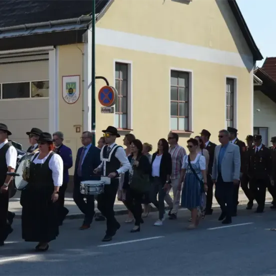 Eine Gruppe von Menschen, einige spielen Trommeln und Trompeten, marschiert in einer Parade die Straße entlang. Hinter ihnen befindet sich ein Gebäude mit einem Schild, das 'Arfang' besagt.