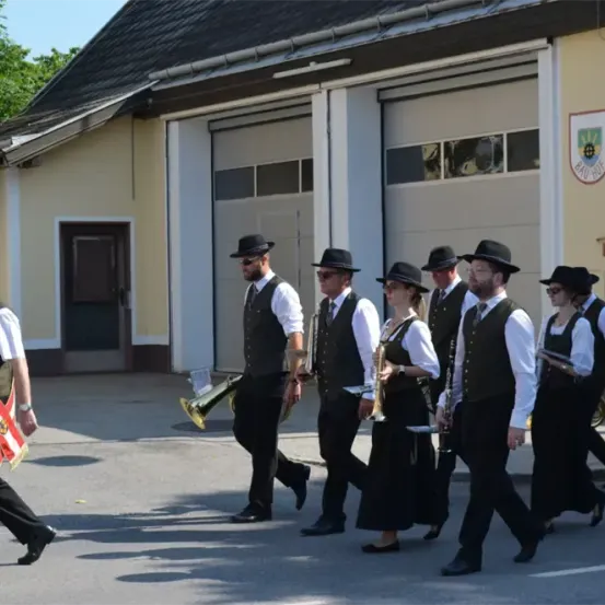 Eine Gruppe von Menschen in traditioneller Kleidung marschiert in einem Umzug. Sie tragen Musikinstrumente und folgen einem Gebäude mit Garagentoren.