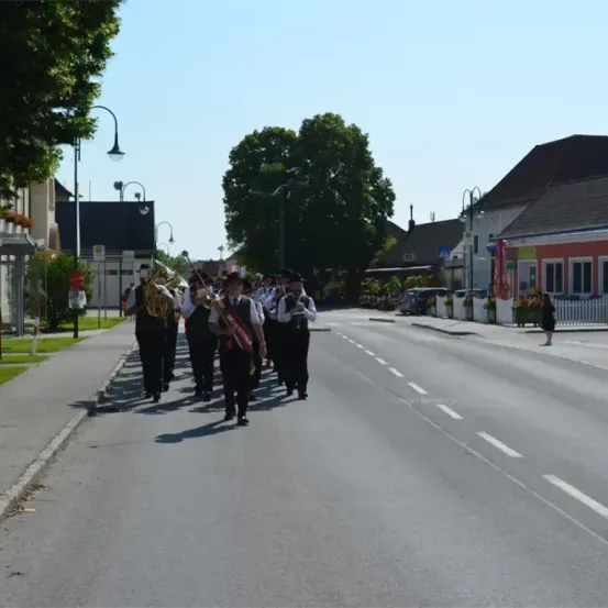 Eine Blaskapelle spielt Instrumente auf einer Straße mit Bäumen und Häusern auf beiden Seiten, wobei eine Person auf dem Bürgersteig geht.