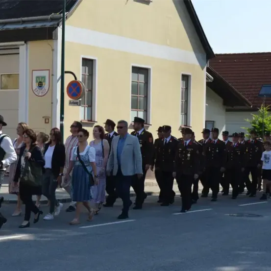 Auf der Straße findet eine Parade statt. In Uniform marschieren mehrere Männer, und vor ihnen sind Zivilisten. Hinter ihnen steht ein Gebäude mit dem Wort 'Bauhof' auf einem Schild.