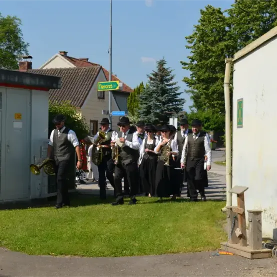 Eine Blaskapelle in traditioneller Kleidung steht auf einer Rasenfläche, möglicherweise in Vorbereitung auf eine Aufführung. Die Bandmitglieder halten Blechblasinstrumente. Im Hintergrund befinden sich Häuser und Bäume.