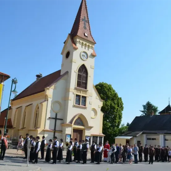 Eine Gruppe von Menschen, möglicherweise eine Blaskapelle, steht vor einer Kirche mit Glockenturm. Die Kirche hat ein braunes Dach und gelbe Wände. Bäume und Gebäude im Hintergrund.