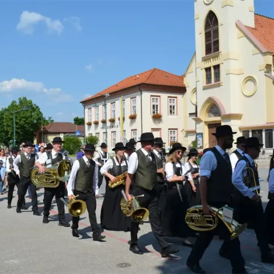 Eine Musikkapelle in traditioneller Kleidung, die Blechblasinstrumente spielt, zieht in einer Parade vor einer Kirche und einem Gebäude mit rotem Dach die Straße entlang.