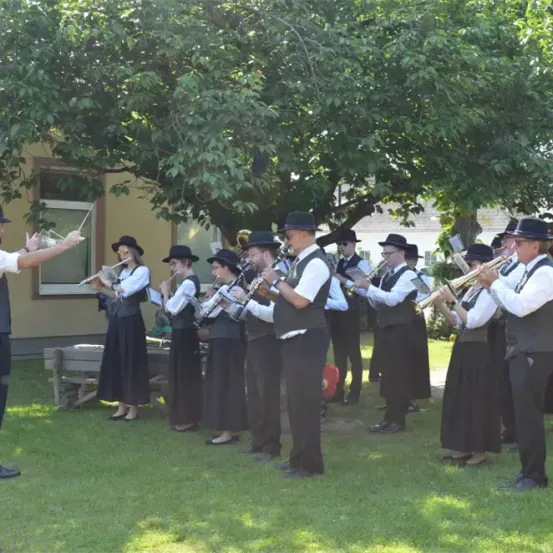 Eine Gruppe von Musikern, die Hüte und schwarze Westen tragen, tritt im Freien auf. Sie spielen verschiedene Instrumente, darunter Flöten und Trompeten. Ein Mann vorne dirigiert die Band. Die Szene spielt unter einem großen Baum, mit einem Gebäude im Hintergrund.