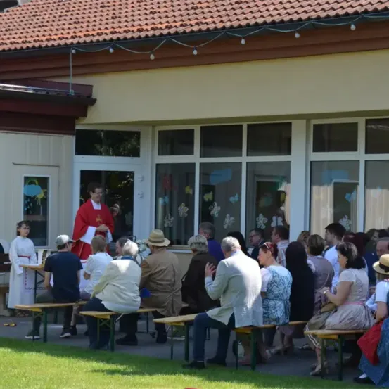 Ein Priester steht vor einer Menschenmenge vor einem Gebäude. Die Leute sitzen auf Bänken und andere stehen. Das Gebäude hat ein rotes Dach und weiße Wände.