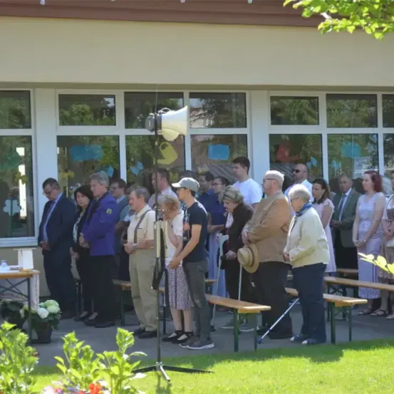 Eine Menschenmenge steht in formeller Kleidung auf einem Bürgersteig vor einem Gebäude. Sie versammeln sich vor einem Gebäude mit Glasfenstern. Ein Mensch mit einem Stock steht davor. Es gibt ein Mikrofon auf einem Stativ. Blumen und Pflanzen sind auf dem Boden.